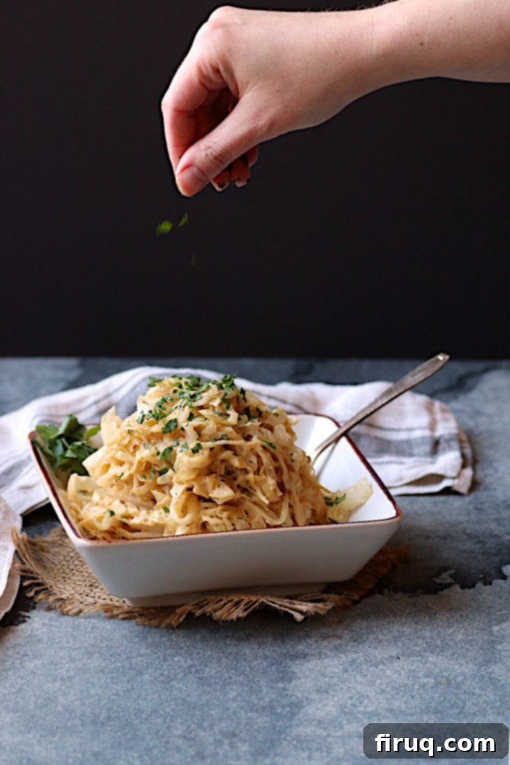 Garnishing Creamy Caramelized Cabbage A hand dropping minced parsley on Creamy Caramelized Cabbage in a square, off-white bowl, adding a final fresh garnish.