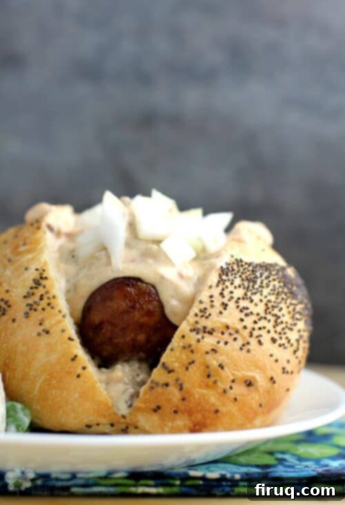 Beer-Glazed Kielbasa and Sausage with Stout Queso 2 front view of the assembled beer brat in a bun with Guinness extra stout queso on top; on a white plate with a grey-blue background