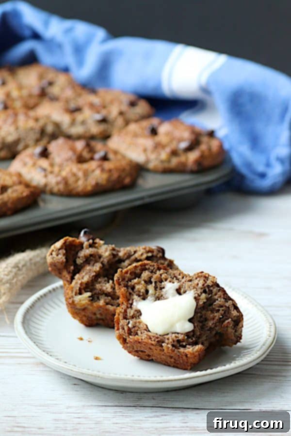 Buttered Banana Chocolate Chip Bran Muffin A single Banana Chocolate Chip Bran Muffin torn in half on a white plate, with one half lightly buttered, showcasing its moist interior.