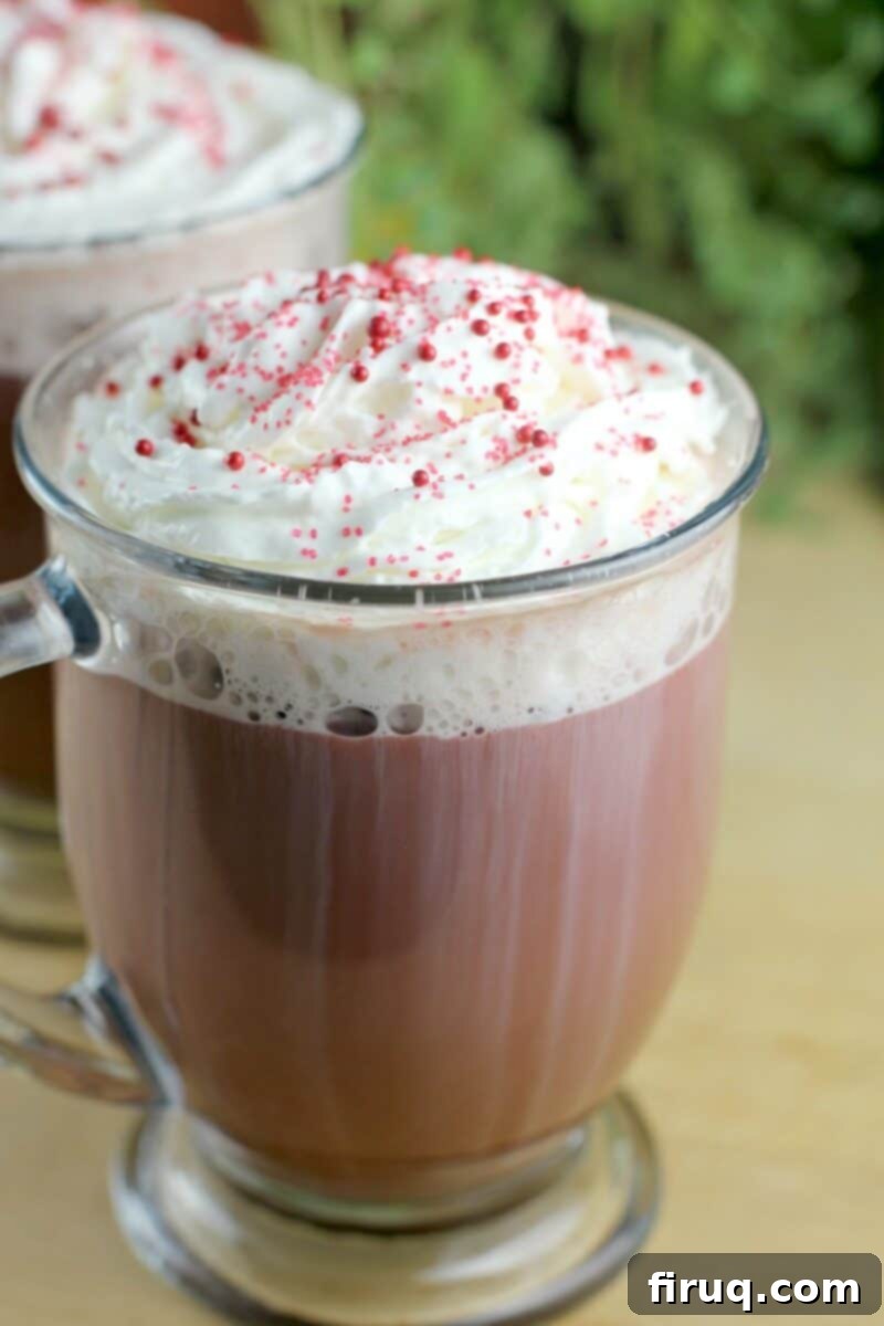 Red Velvet Latte 2 Red Velvet Latte served in a clear glass mug on a rustic wooden board, with a second blurry mug and green plant leaves in the background.