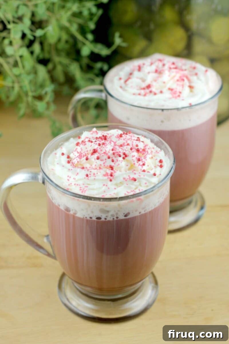 Red Velvet Latte 4 Two glass mugs of Red Velvet Latte, generously topped with whipped cream, arranged on a wooden board. A vibrant green plant is visible in the soft-focus background.