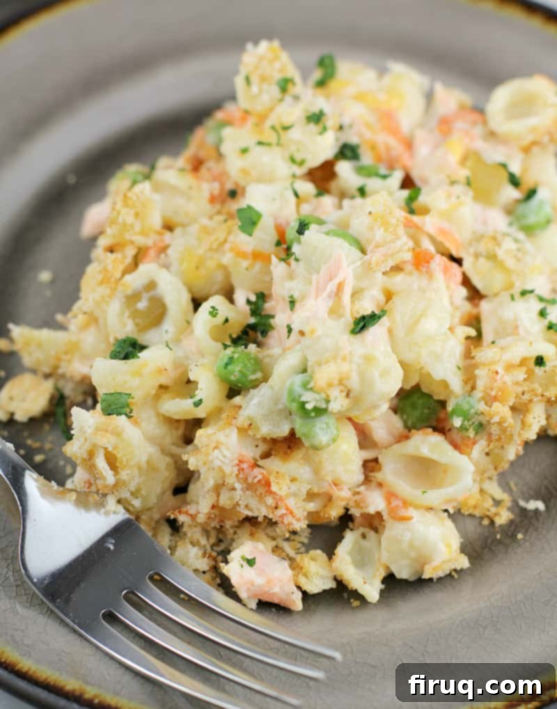 Golden Salmon Casserole 3 Top-down view of a serving of fresh salmon casserole on a white plate with a fork, showing the creamy pasta, salmon, and cracker topping