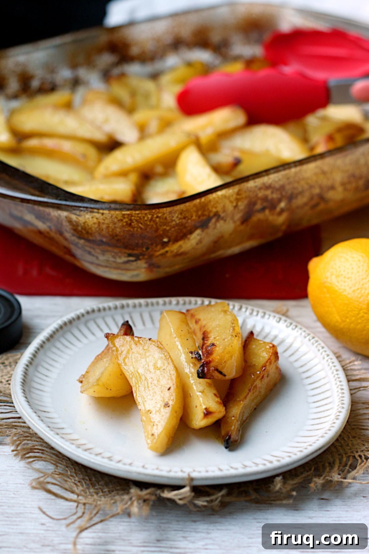 Serving Greek Lemon Potatoes Greek Lemon Potatoes on a small white plate with a glass baking dish full of the same in the background.