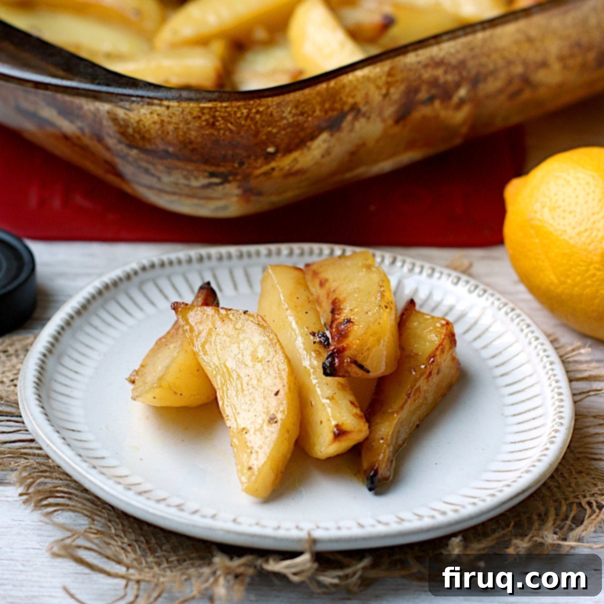 Perfectly Roasted Greek Lemon Potatoes Greek Lemon Potatoes on a small white plate with a glass baking dish full of the same in the background.