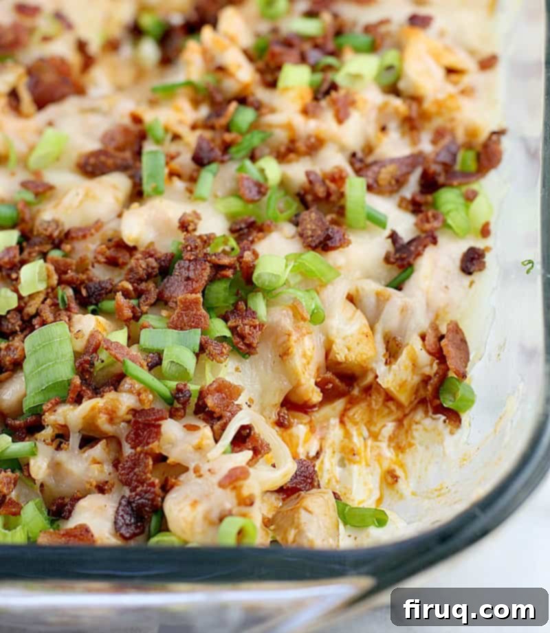 Loaded Buffalo Bacon Chicken Dip 6 Overhead view of the buffalo bacon chicken dip in a clear baking dish, with a section scooped out to reveal the distinct layers of creamy ranch-cheese, spicy buffalo chicken, and melted cheese, topped with crispy bacon and fresh green onions.