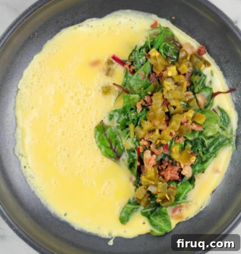 Keto Dairy-Free Bacon Jalapeno Popper Omelet 4 Top down view of omelet toppings (greens, bacon, and jalapeno) on egg cooking in skillet.