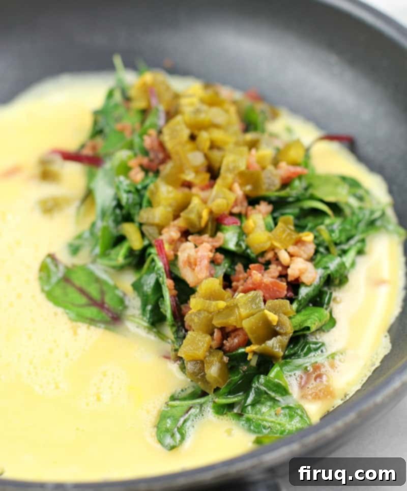 Keto Dairy-Free Bacon Jalapeno Popper Omelet 5 Side view of omelet toppings (greens, bacon, and jalapeno) on egg cooking in skillet.