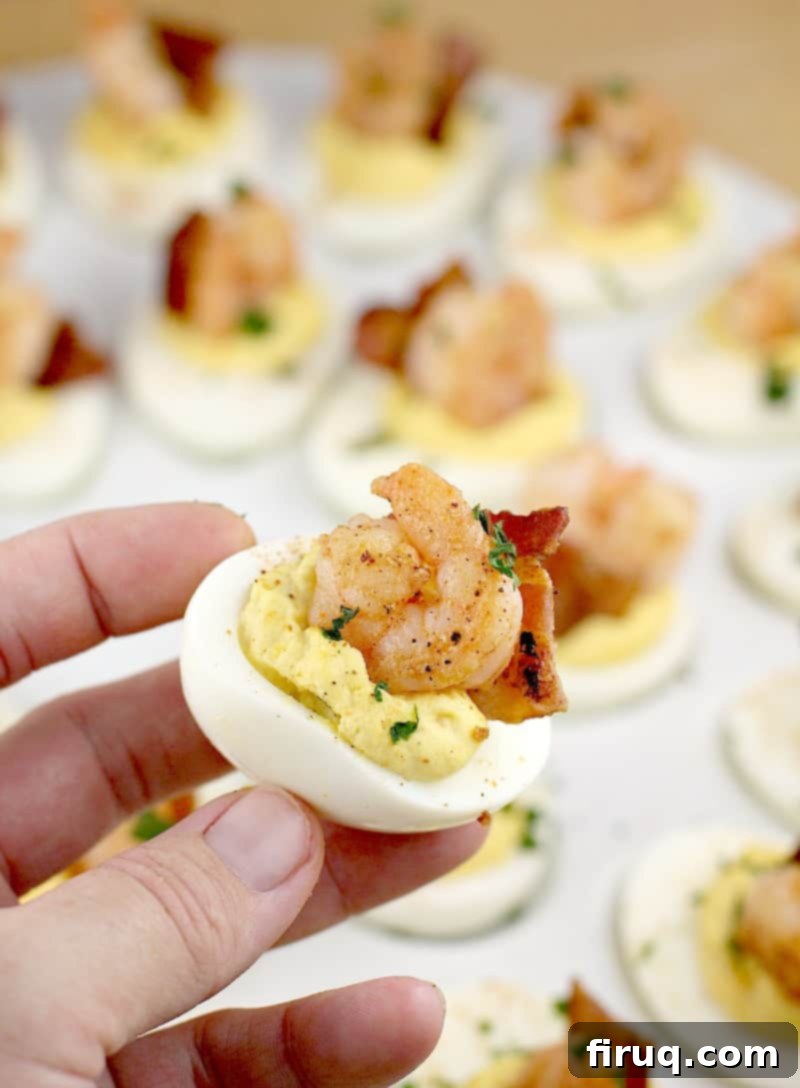 Zesty Creole Shrimp Deviled Eggs with Crispy Bacon 3 a hand holding up one deviled egg with bacon and shrimp; a white platter of others is in the background, showing the rich filling and garnish