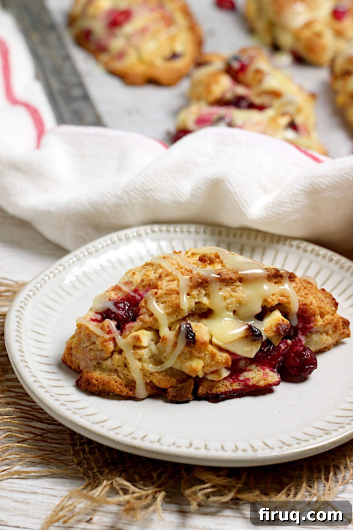 Closeup of Cranberry Orange Scone A closeup photo of a Cranberry Orange Scone with White Chocolate on a small white plate, showing the flaky texture and visible cranberries and white chocolate.