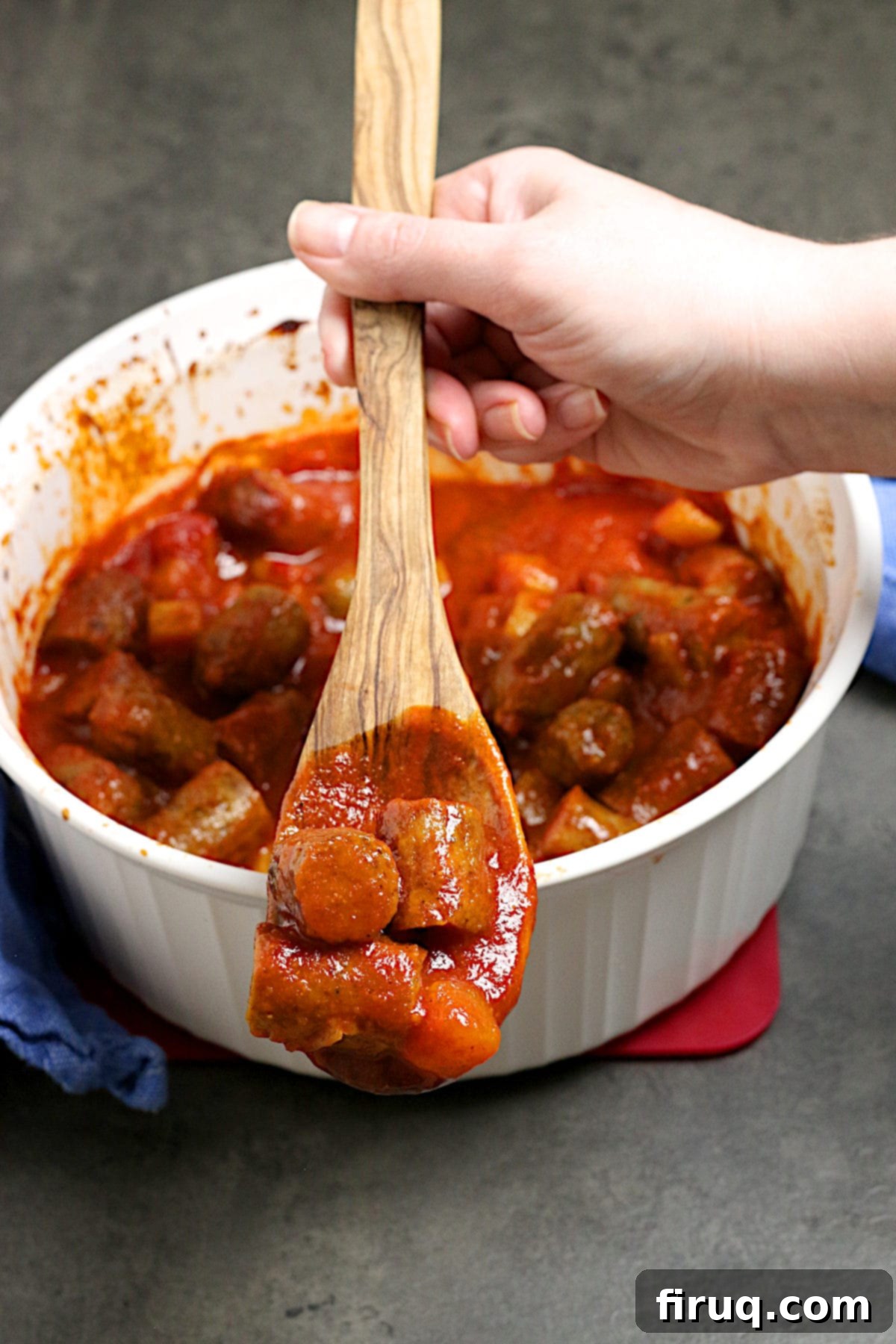Serving Peachy Sweet and Sour Sausages A hand scooping Peachy Sweet and Sour Sausages from a white casserole dish on a wooden spoon.