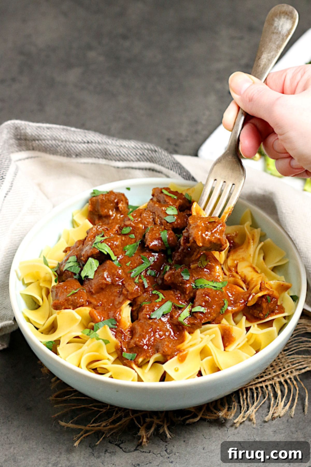 Hearty Beef Paprika with Egg Noodles A fork scooping some Beef Paprika and egg noodles from a pale blue bowl.