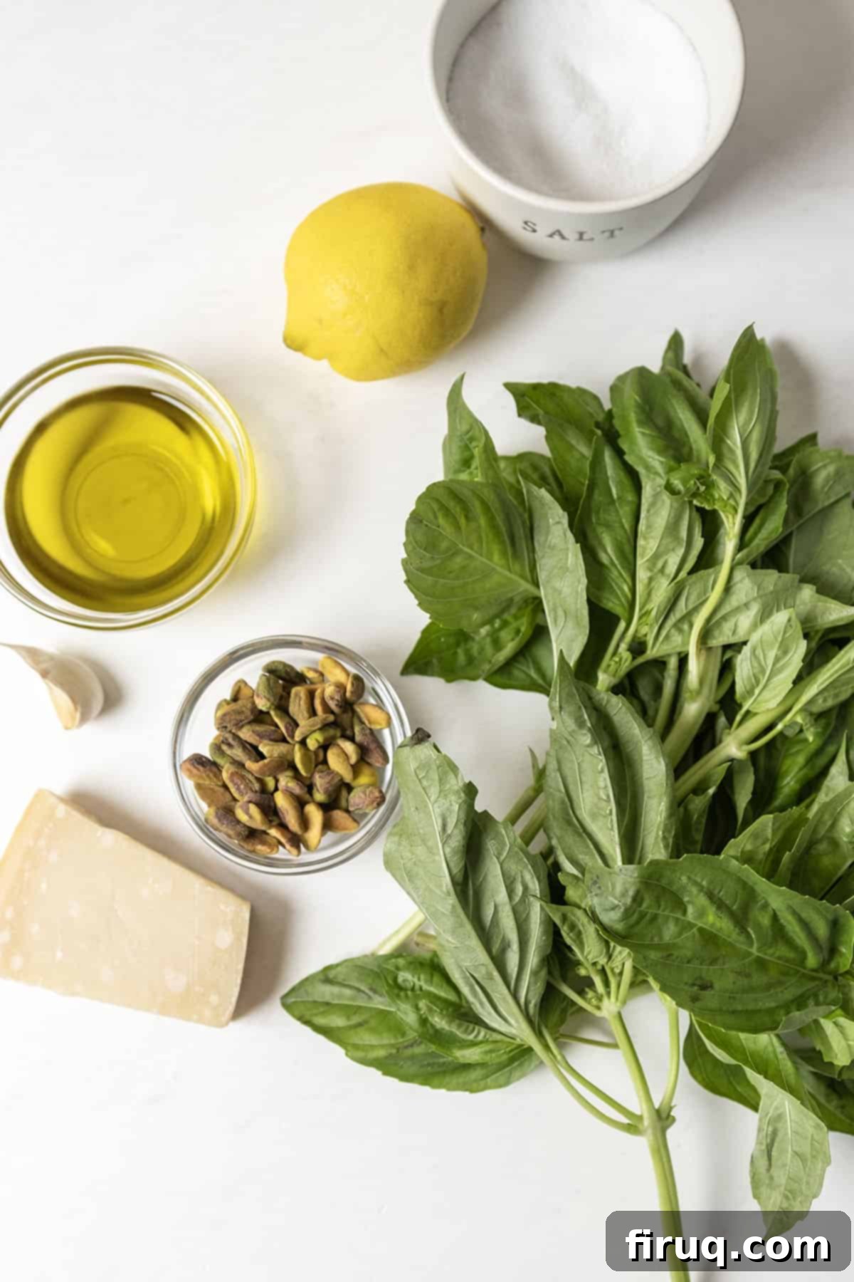 Emerald Pistachio Pesto 3 A flat lay of fresh basil leaves, shelled pistachios, a head of garlic, a lemon, a block of Parmigiano-Reggiano cheese, and a bottle of olive oil, which are the main ingredients for pistachio pesto.