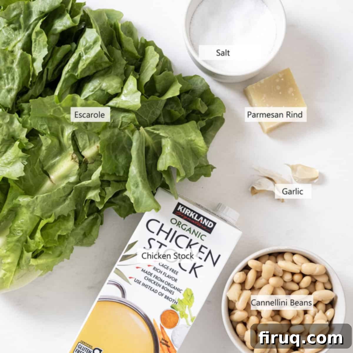 Hearty Escarole and Bean Soup 3 A vibrant flat lay of fresh ingredients for Escarole and Bean Soup, including a head of broad-leafed escarole, a can of cannellini beans, garlic cloves, a parmesan rind, and olive oil, all neatly arranged.