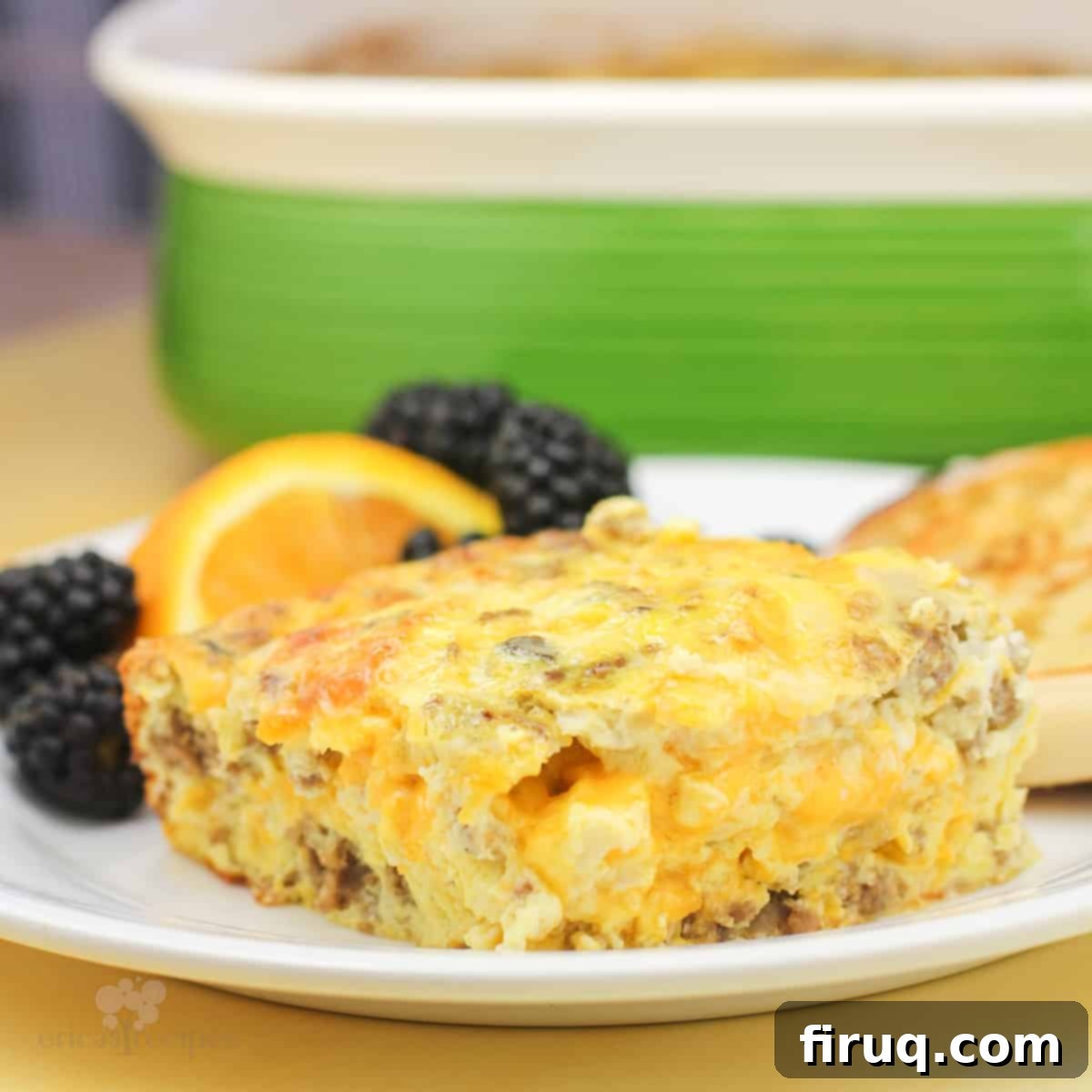 Hearty Sausage & Vegetable Bake 2 A golden-brown sausage egg and cheese casserole portion on a white plate with fresh fruit garnish, set against a blurred background of a green casserole dish.