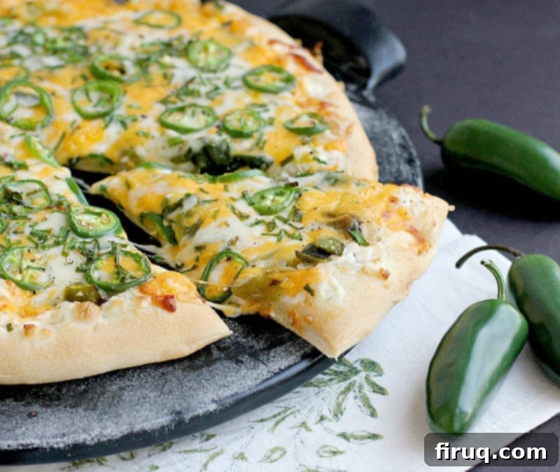 Popper's Paradise Pie 2 side view of the finished jalapeno pizza on a black pizza stone with one slice cut and nudged out a bit to show melty cheese. a white napkin with green stencil and fresh jalapenos are placed as decoration