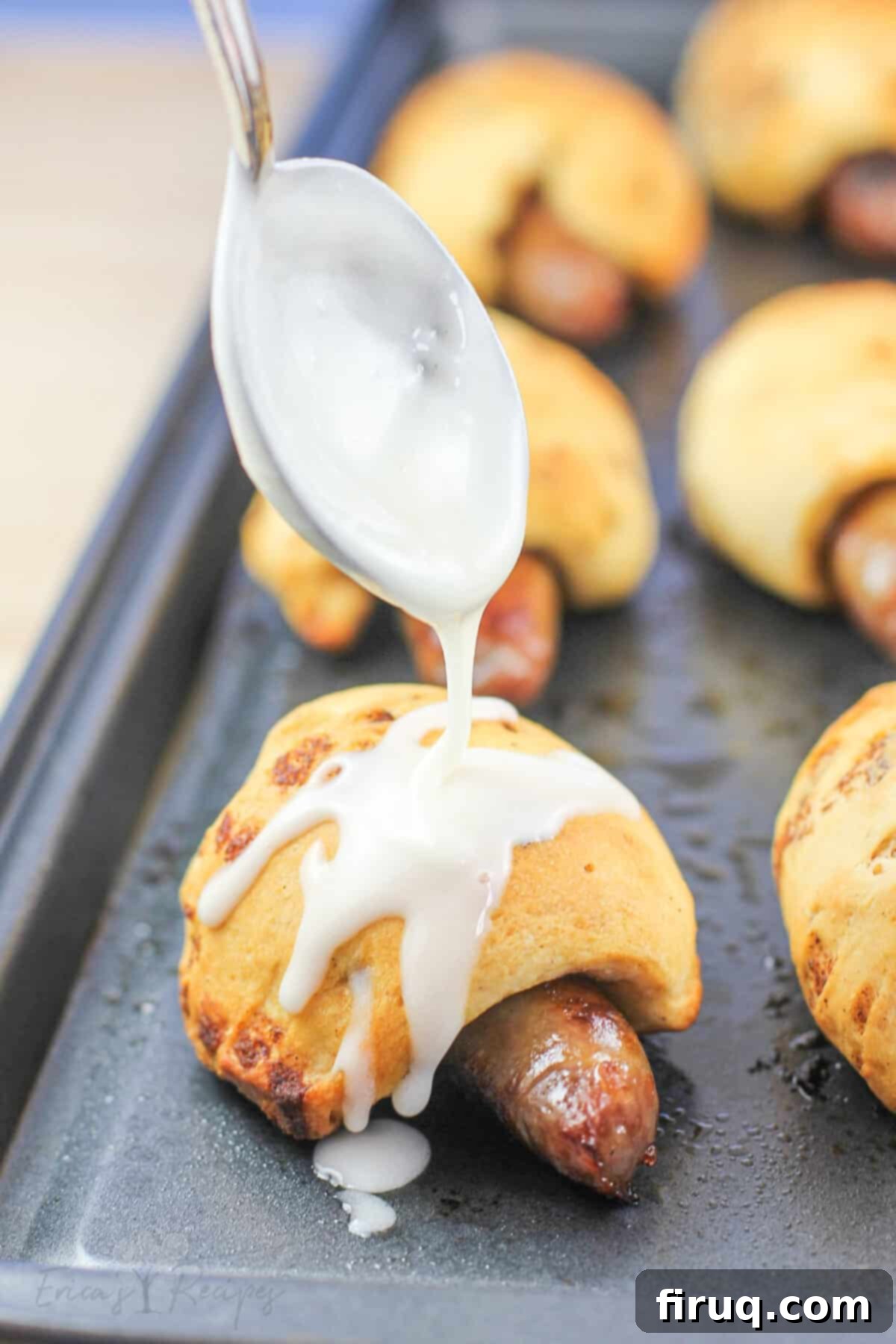 Cinnamon Swirl Sausage Bites 2 Cinnamon Roll Pigs in a Blanket on a metal baking sheet, with a spoon drizzling maple icing over them.