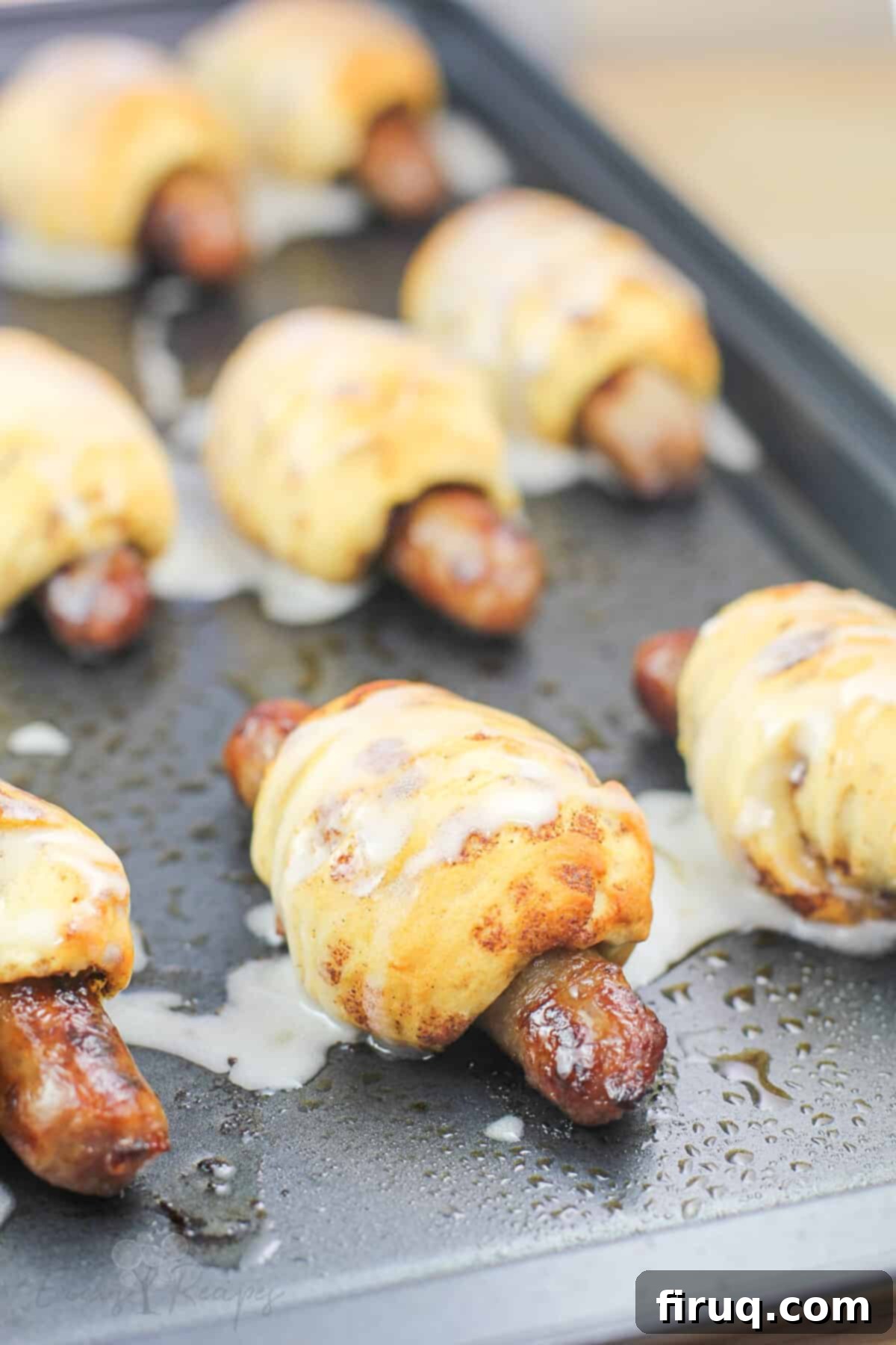Cinnamon Swirl Sausage Bites 8 Baked Cinnamon Roll Pigs in a Blanket on a metal baking sheet, generously topped with maple icing.