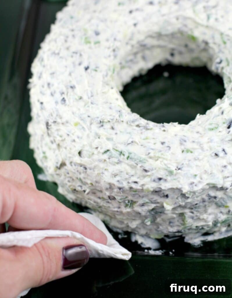 Festive Olive & Scallion Wreath Dip 10 A hand using a paper towel to carefully clean smudges from the plate around the base of the newly formed Christmas cheese wreath, ensuring a pristine presentation.