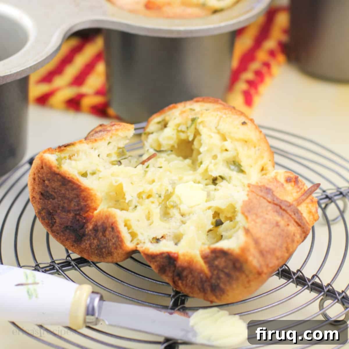 Poblano Cloud Bites 2 A perfectly split poblano pepper popover resting on a wire rack with a small butter knife, a popover pan subtly blurred in the background, highlighting its crispy texture and hollow interior.