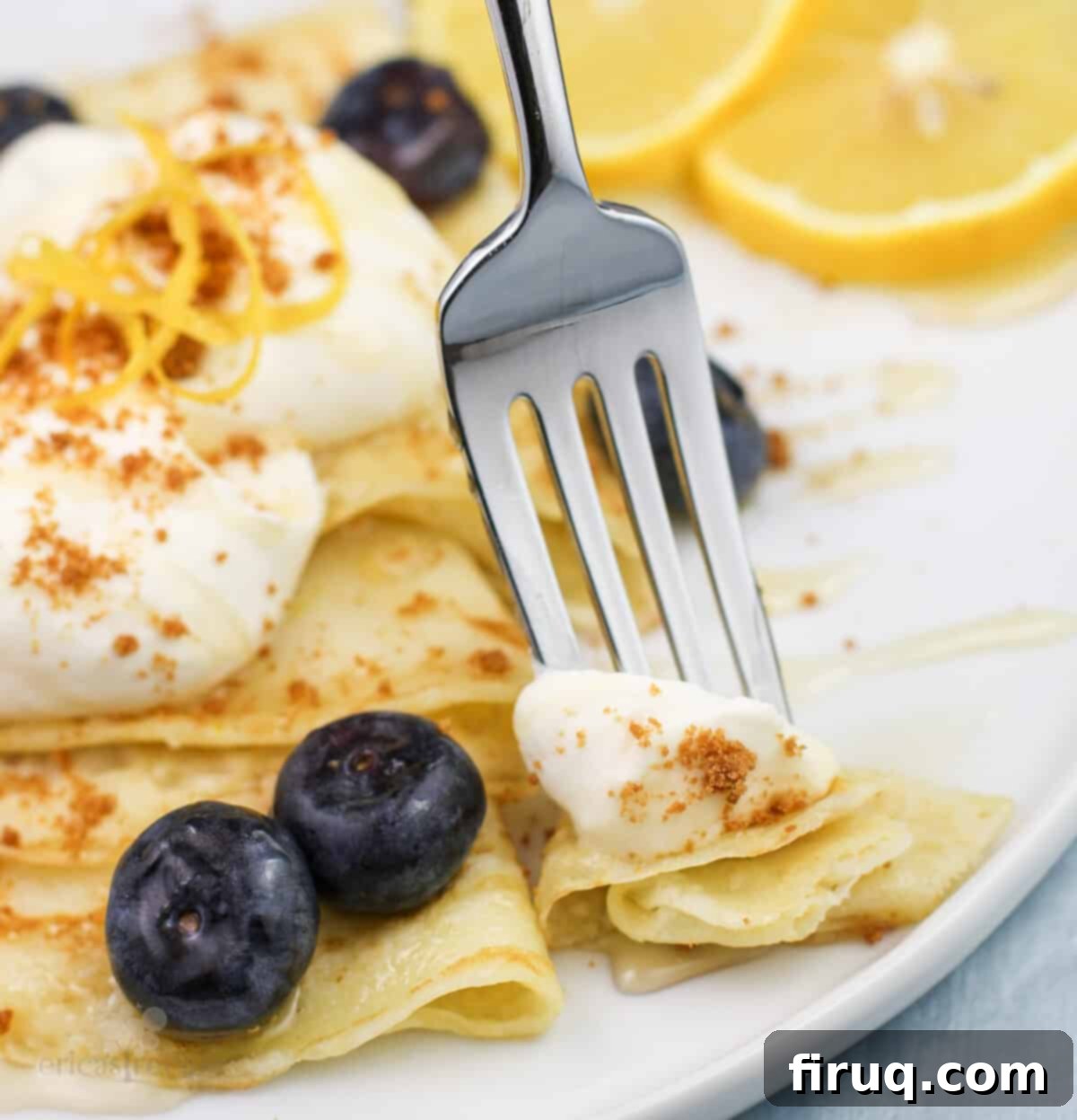 Zesty Lemon Crepes with Cloud Mascarpone 2 Fork lifting a bite of lemon crepe with mascarpone, blueberries, and honey on a white plate