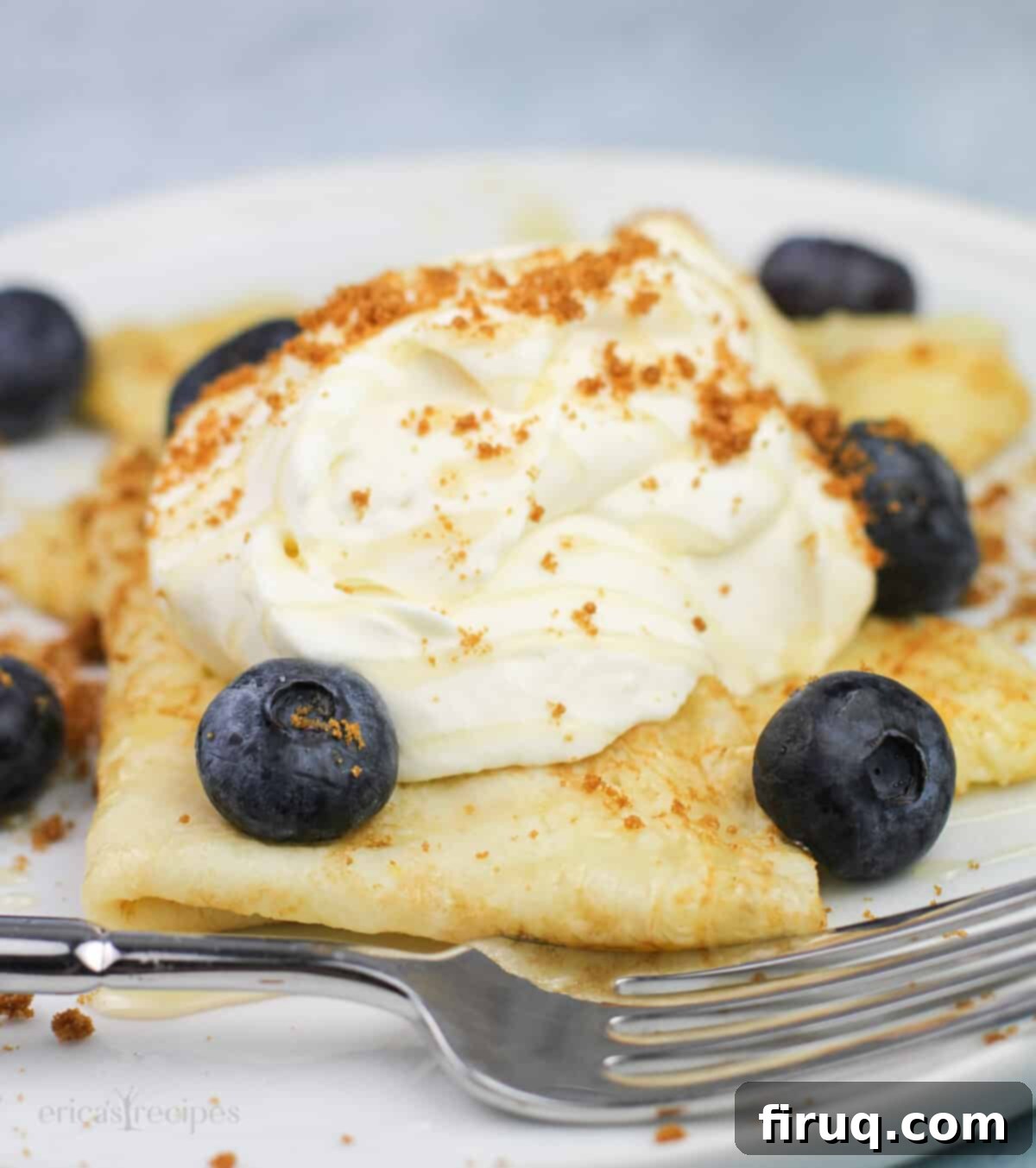 Zesty Lemon Crepes with Cloud Mascarpone 4 A single serving of elegant lemon crepes with lemon mascarpone, blueberries, honey, and crushed Biscoff cookies on a white plate, ready to be enjoyed with a fork