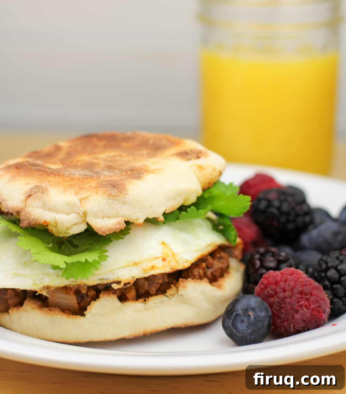 Taco Morning Stack 3 A freshly prepared breakfast sandwich featuring taco meat, egg, and cheese on an English muffin, neatly arranged on a white plate next to colorful berries.